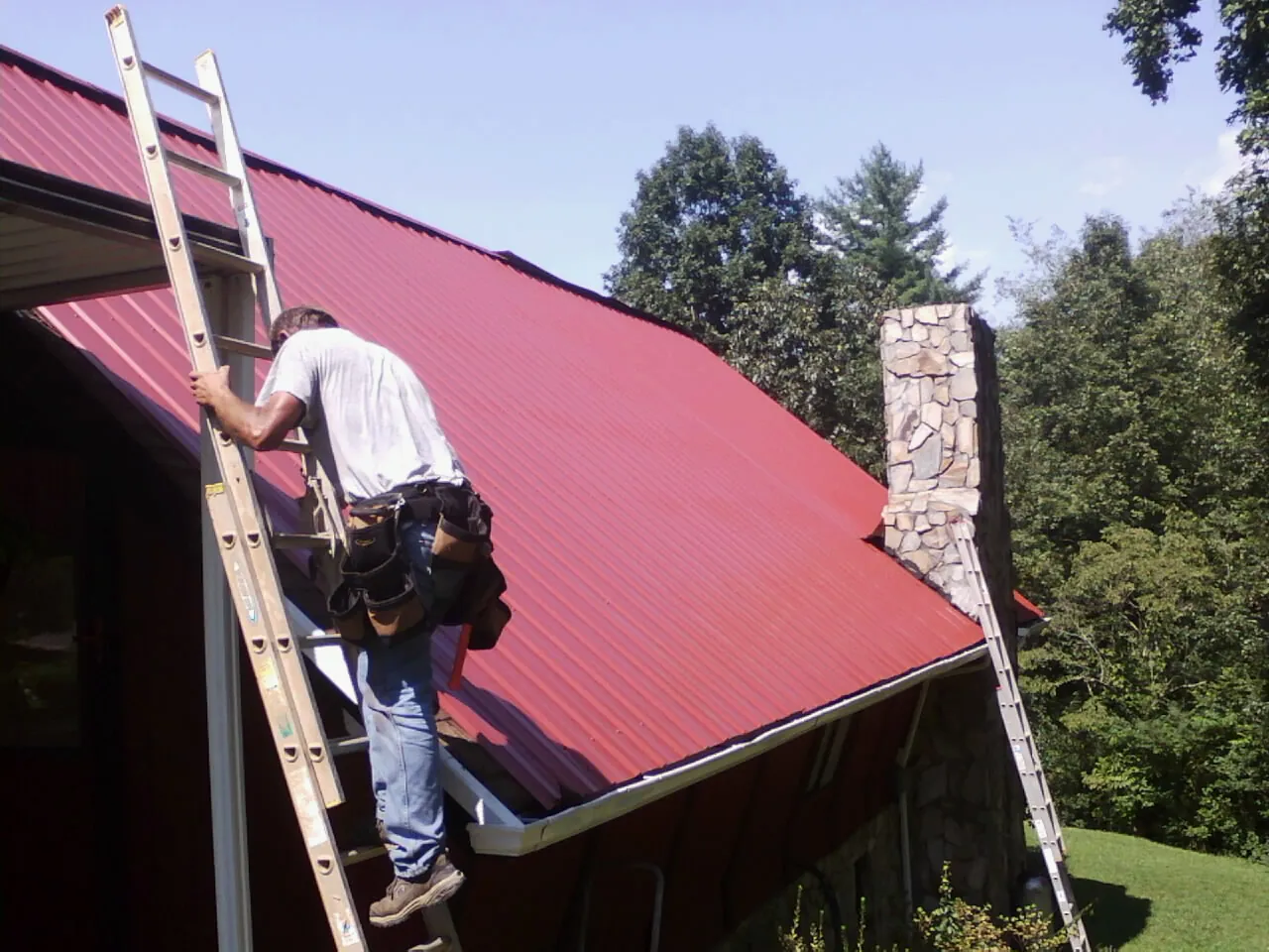 Roofer on a ladder beside a red metal roof and stone chimney — Soffit & Fascia Repair by Roofing Brothers in Conestoga